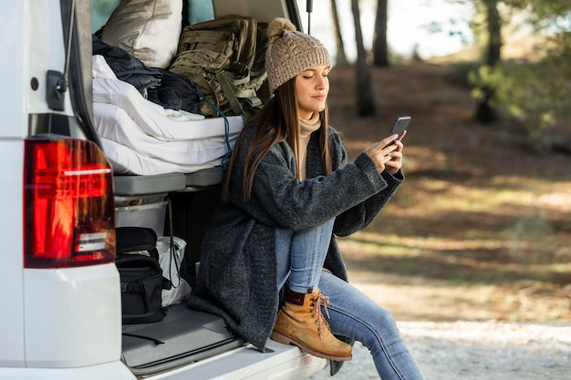 Vue latérale d'une femme assise dans le coffre de la voiture lors d'un voyage sur la route et à l'aide de smartphone