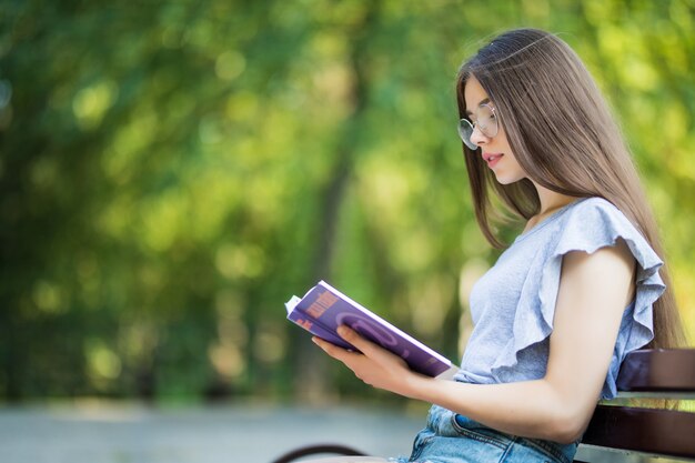 Vue latérale d'une femme brune heureuse à lunettes assis sur un banc et livre de lecture dans le parc