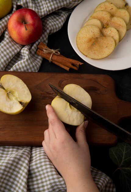 Vue latérale femme coupe la pomme jaune sur une planche avec des tranches de cannelle et de pomme sur une plaque sur une nappe à carreaux