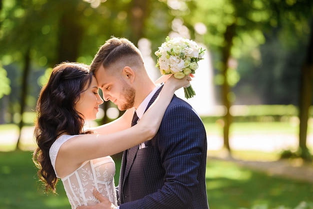 Vue latérale d'une femme heureuse avec des cheveux bouclés et un beau maquillage portant une robe à la mode étreignant le marié par le cou