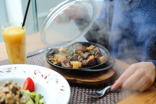 Photo gratuite vue latérale une femme a ouvert le couvercle pommes de terre frites avec un foie dans une poêle sur un support avec du jus d'orange sur la table