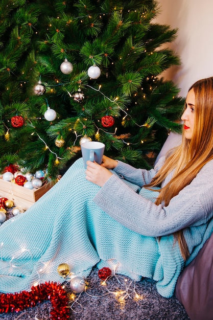 Photo gratuite vue latérale d'une femme tenant une tasse en face de l'arbre de noël