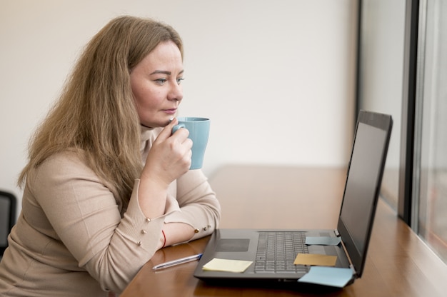 Vue latérale d'une femme tenant une tasse et travaillant sur un ordinateur portable au bureau