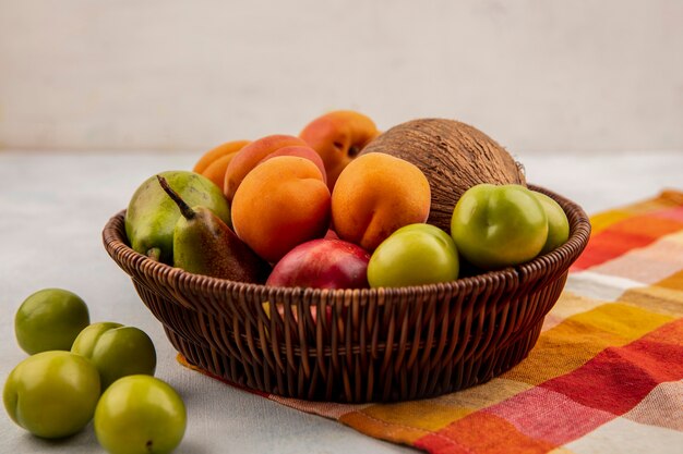 Vue latérale des fruits comme poire pêche abricot noix de coco dans le panier sur tissu à carreaux avec des prunes sur fond blanc