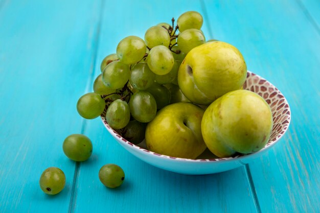 Vue latérale des fruits comme le raisin et les pluots verts dans un bol et des baies de raisin sur fond bleu