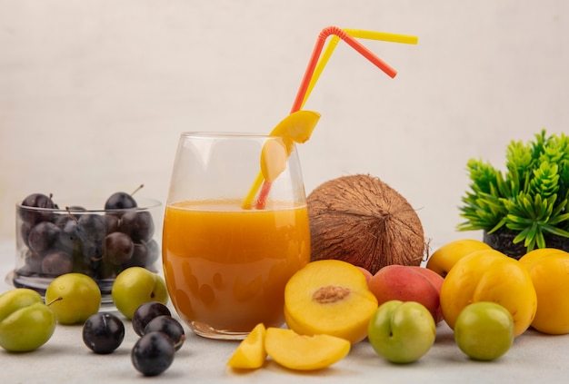 Vue latérale des fruits sains et colorés tels que les prunelles sur un bol en verrepeachescoconut avec du jus de pêche frais sur un verre sur un fond blanc