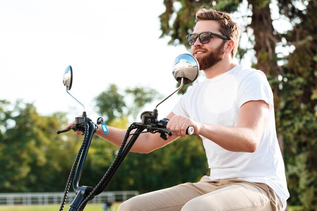 Vue latérale de gai homme berded en lunettes de soleil promenades en moto moderne à l'extérieur et à l'écart