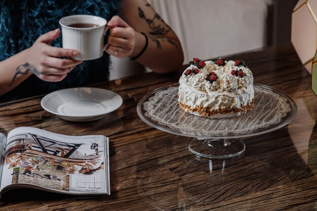 Vue latérale d'un gâteau avec de la crème fouettée et des baies sur un support en verre et une personne buvant du thé à la table