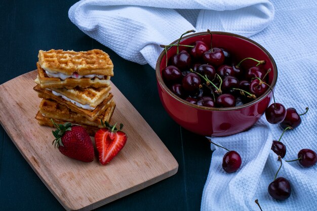 Vue latérale des gâteaux et des fraises sur une planche à découper avec des cerises dans un bol sur un tissu sur fond noir