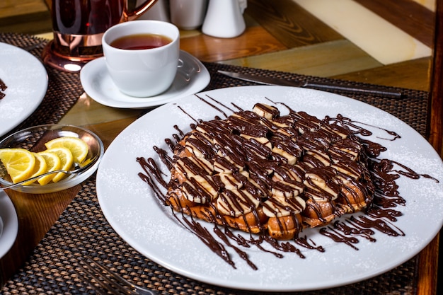 Vue latérale de la gaufre aux bananes recouvertes de chocolat sur une plaque blanche servie avec du thé sur la table