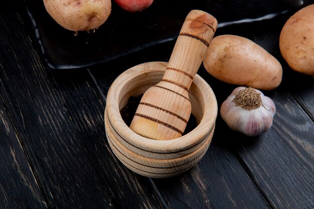 Photo gratuite vue latérale des graines de poivre noir dans un broyeur d'ail avec de l'ail et des pommes de terre sur une surface en bois