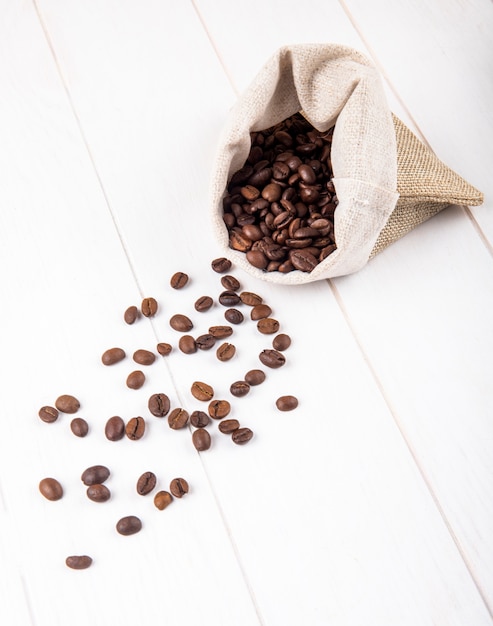Photo gratuite vue latérale des grains de café éparpillés dans un sac sur fond de bois blanc