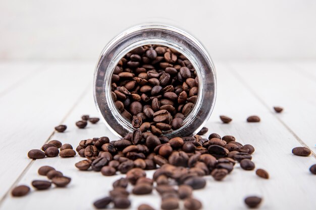 Vue latérale des grains de café frais tombant d'un bocal en verre sur un fond en bois blanc