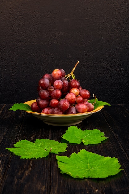 Vue latérale d'une grappe de raisin doux dans une assiette et des feuilles de vigne verte sur une table rustique