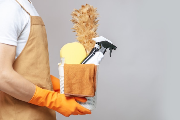 Vue latérale, gros plan sur la main du jeune homme en tablier et gants en caoutchouc tenant un panier de matériel de nettoyage, le plumeau, le vaporisateur, l'éponge et le chiffon pour essuyer dans le panier