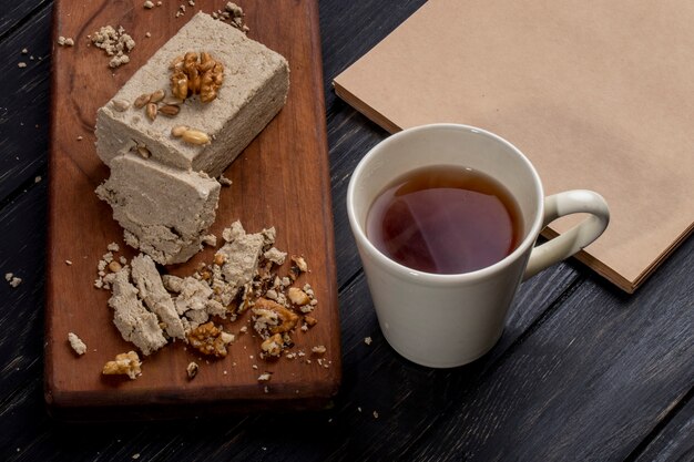 Vue latérale de halva avec des graines de tournesol et des noix sur une planche de bois et une tasse de thé sur rustique