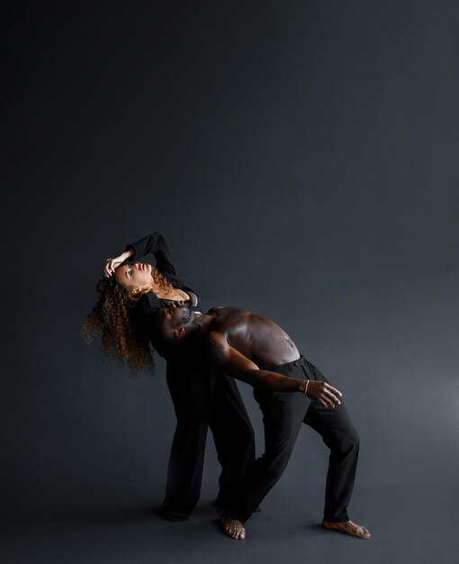 Vue latérale d'un homme afro-américain aux seins nus et d'une femme aux cheveux bouclés en costume pliant les corps en arrière tout en répétant le mouvement de danse dans un studio noir isolé