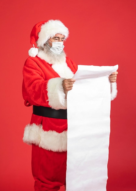 Vue latérale de l'homme en costume de père Noël avec masque médical tenant la liste des cadeaux