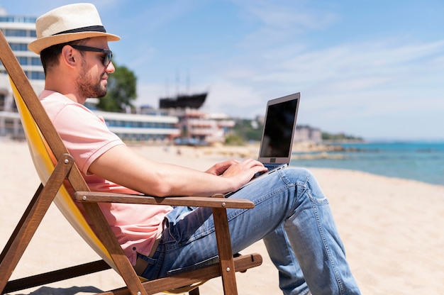 Vue latérale de l'homme dans une chaise de plage travaillant sur ordinateur portable