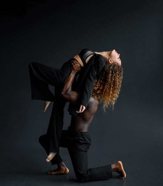 Vue latérale d'un homme fort à la peau sombre en pantalon noir appuyé sur le genou tout en soulevant une femme sensuelle en costume noir et lingerie lacée tout en répétant la danse sur fond noir