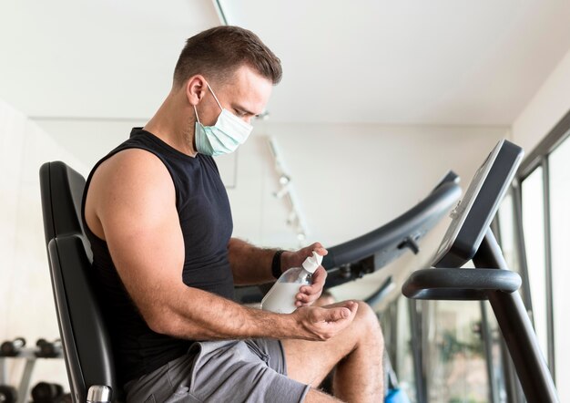 Vue latérale de l'homme avec un masque médical à l'aide d'un désinfectant pour les mains au gymnase