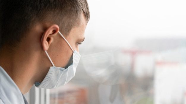 Vue latérale de l'homme avec un masque médical regardant par la fenêtre