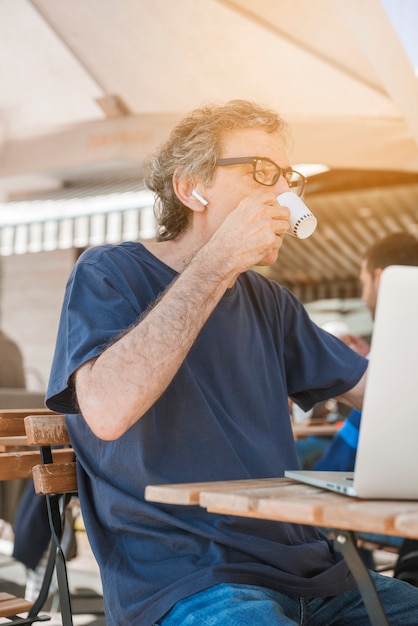 Vue latérale d&#39;un homme senior avec ordinateur portable, boire du café au café en plein air