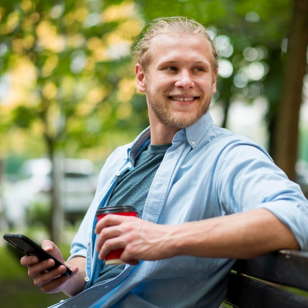 Vue latérale de l'homme souriant à l'extérieur avec smartphone et tasse