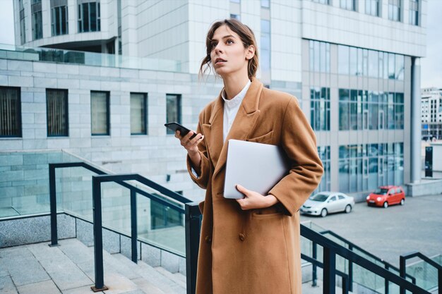 Vue latérale d'une jeune femme d'affaires séduisante en manteau avec ordinateur portable et téléphone portable regardant pensivement à l'extérieur