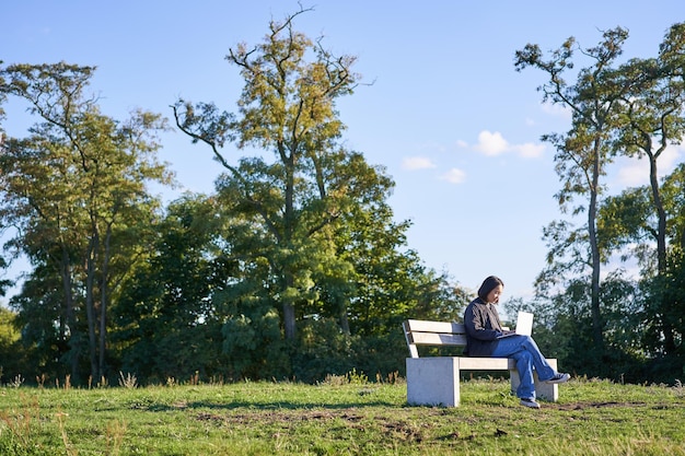 Photo gratuite vue latérale d'une jeune femme assise seule dans un parc sur un banc à l'aide de son ordinateur portable pour étudier ou travailler à distance f