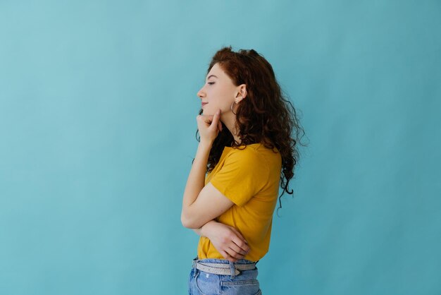 Vue latérale d'une jeune femme rousse souriante en chandail jaune posant isolé sur fond bleu turquoise en studio Concept de mode de vie des gens Tenir les mains croisées en regardant de côté