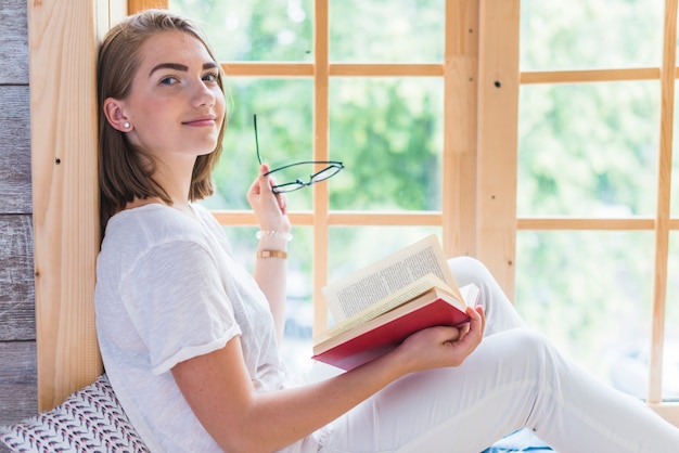 Photo gratuite vue latérale de la jeune femme tenant des lunettes et un livre près de la fenêtre