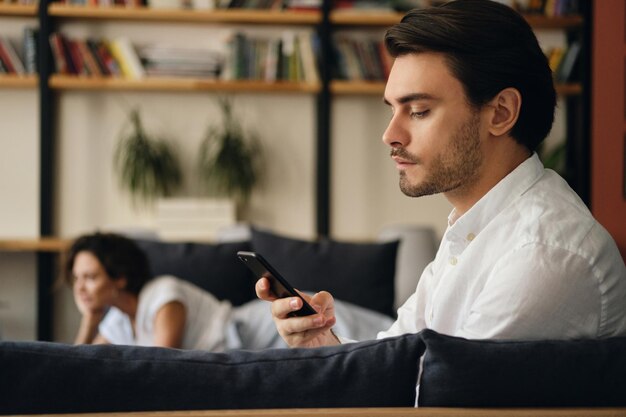 Vue latérale d'un jeune homme d'affaires beau assis sur un canapé avec un collègue en arrière-plan tout en utilisant judicieusement un téléphone portable dans un espace de coworking moderne