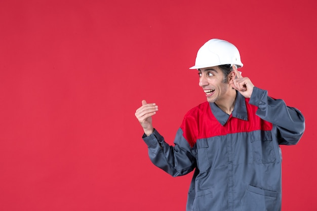 Vue latérale d'un jeune travailleur confus en uniforme avec un casque pointant vers le haut et regardant le côté droit sur un mur rouge isolé