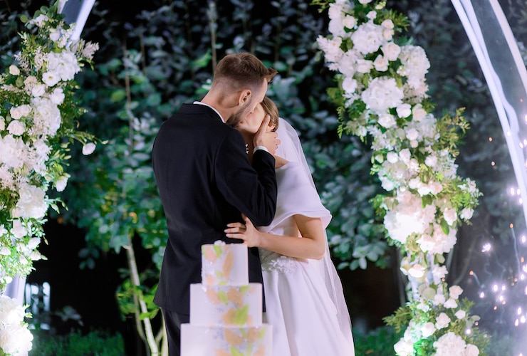 Photo gratuit vue latérale des jeunes mariés s'embrassant pendant la célébration du mariage sur fond de décor avec des fleurs