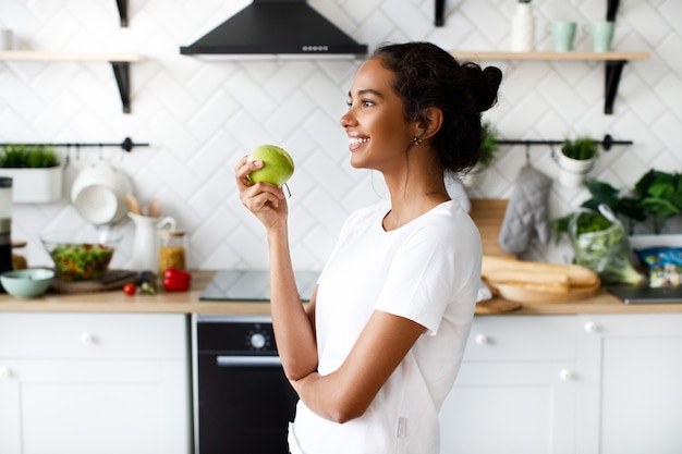 Vue latérale d'une jolie femme mulâtre souriante qui tient une pomme et regarde loin sur la cuisine moderne blanche