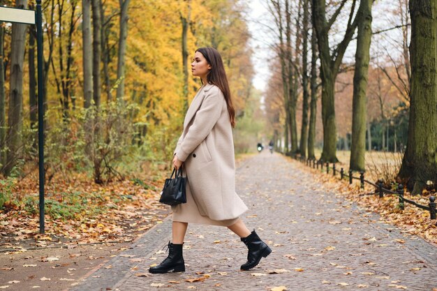 Vue latérale d'une jolie fille brune en manteau marchant seule dans le magnifique parc d'automne
