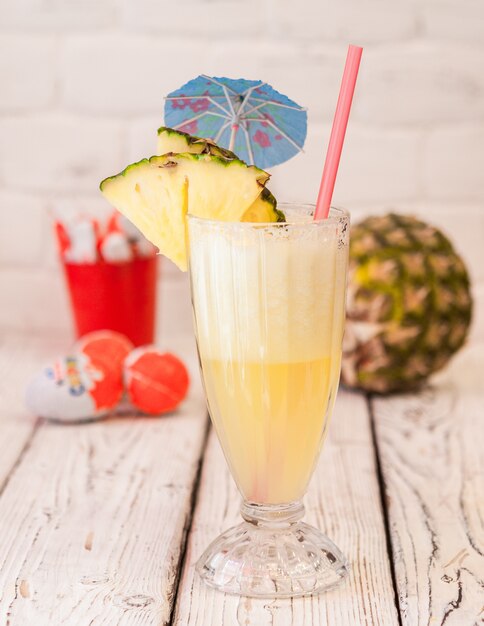 Vue latérale jus d'ananas avec jus de paille et tranches d'ananas et parapluie cocktail dans une tasse en verre sur une table en bois