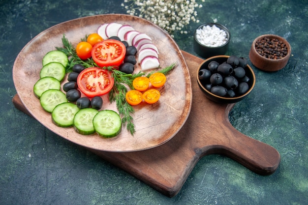 Vue latérale de légumes frais hachés dans une assiette brune sur une planche à découper en bois olives dans un bol fleur d'ail sel sur fond de couleurs mélangées