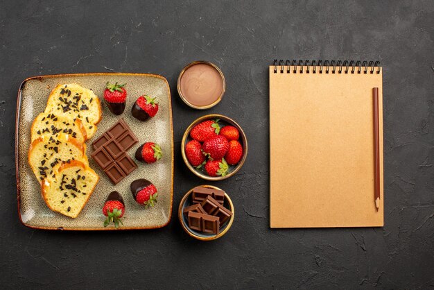 Vue latérale de loin tasse de thé avec gâteau assiette de gâteau avec fraises enrobées de chocolat tasse de thé crème au chocolat et fraises dans des bols à côté d'un crayon et d'un cahier