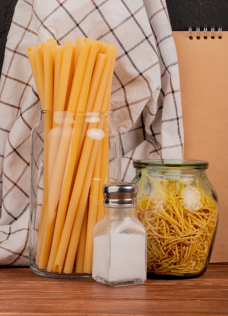 Vue latérale des macaronis comme bucatini et spaghetti avec du sel et un tissu à carreaux et bloc-notes sur la surface en bois