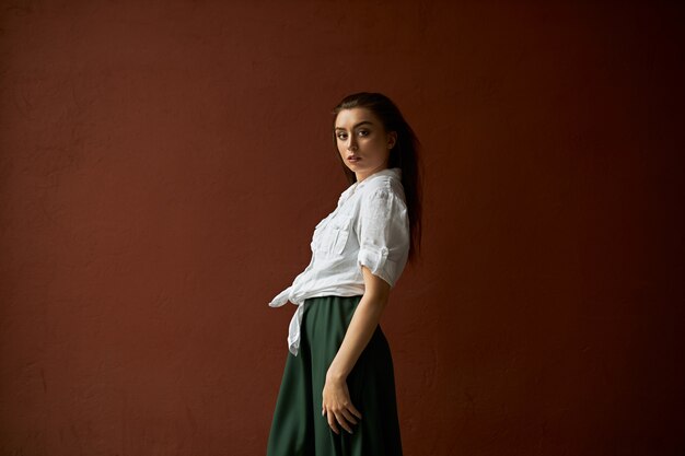 Vue latérale de la magnifique jeune femme mince en chemise blanche et jupe posant contre fond de mur de studio de copie espace pour votre publicité ayant une expression faciale confiante, regardant la caméra