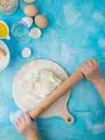 Photo gratuite vue latérale de la main de femme, pétrir la pâte sur une planche à rouler avec un rouleau à pâtisserie et des œufs d'eau sur fond bleu avec copie espace