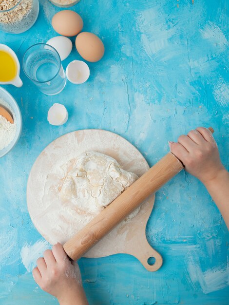 Vue latérale de la main de femme, pétrir la pâte sur une planche à rouler avec un rouleau à pâtisserie et des œufs d'eau sur fond bleu avec copie espace