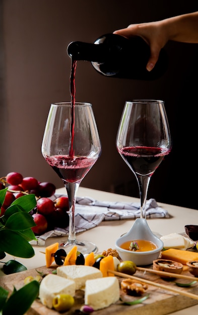 Vue latérale de la main de femme versant du vin rouge dans du verre et différents types de fromage raisin olive noyer sur la surface blanche et fond noir