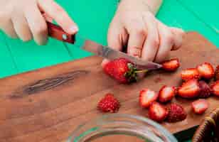 Photo gratuite vue latérale des mains coupant des fraises avec un couteau sur une planche à découper sur la surface verte