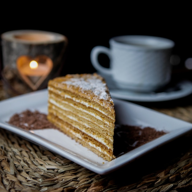Photo gratuite vue latérale d'un morceau de gâteau au miel avec une tasse de thé et une bougie et une assiette blanche dans des serviettes de service