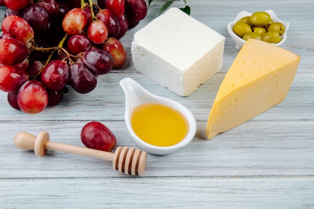 Vue latérale de morceaux de fromage avec du miel, du raisin frais et des olives marinées sur une table en bois gris