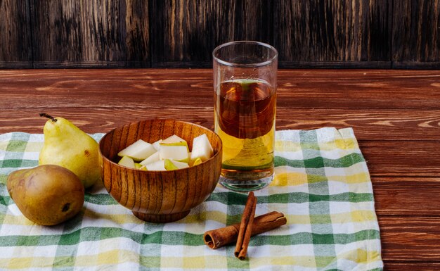 Vue latérale de morceaux de poire dans un bol en bois avec un verre de limonade et des bâtons de cannelle sur une nappe à carreaux