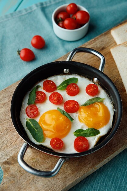 Vue latérale des œufs au plat avec des tomates dans une poêle décorée de feuilles avec des tranches de pain sur une planche à découper sur fond bleu